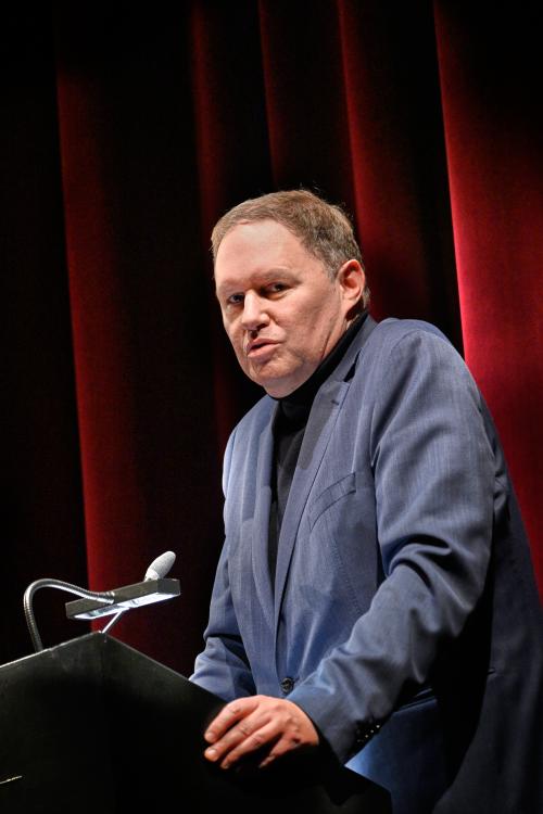 Kultursenator Dr. Carsten Brosda in einem blauen Jackett an einem schwarzen Stehpult. Er schaut zur Seite, sein Mund ist geöffnet. Im Hintergrund ist ein geschlossener roter Samtvorhang zu sehen.