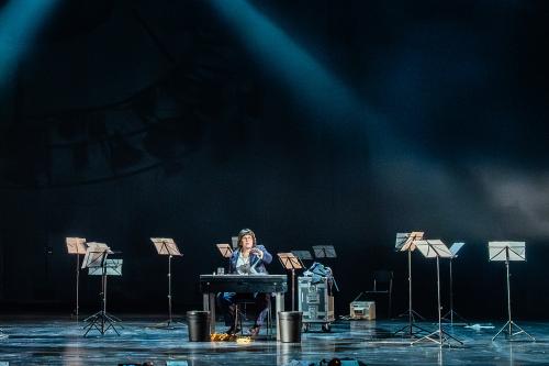 Julia Wieninger sitzt in einem lila-grün-karierten Anzug mit weißem Hemd an einem Tisch. Neben dem Tisch stehen zwei schwarze Mülleimer. Ihre linke Hand ist erhoben, ihr Mund ist geöffnet. Um sie herum stehen viele Notenständer quer im Raum verteilt.