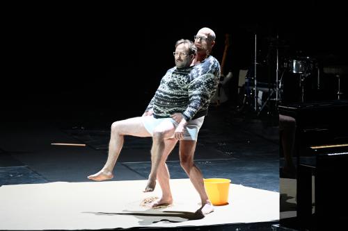 Bühnensituation: Joachim Meyerhoff und Samuel Weiss stecken gemeinsam in einem großen grauen Norwegerpullover, Weiss befindet sich vor Meyerhoff, er ist etwas kleiner als er. Beide tragen weiße Unterhosen, ihre Beine und Füße sind nackt. Joachim Meyerhoff lehnt sich etwas zurück, sodass die Beine von Samuel Weiss angehoben sind, er hält sie angewinkelt in der Luft. Hinter ihnen steht ein gelber Plastikeimer, auf dem Boden daneben ein kleiner Haufen Sand.