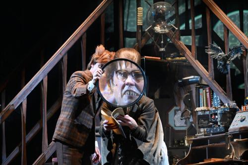 Jan-Peter Kampwirth und Ute Hannig stehen vor zwei gegenüber zulaufenden Treppen in einem eingerichteten Büro, mit vielen wissenschaftlichen Instrumenten. Jan-Peter hält eine überdimensionale Lupe vor Utes Gesicht, wodurch man es vergrößert sieht.