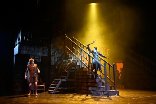 Ute Hannig steht auf einer Treppe eines großen Bücherregals und streckt eine lange Axt in die Höhe. Jan-Peter Kampwirth guckt sie von unten an. 