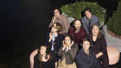 Das Ensemble von „Die Möwe“ sitzt verstreut auf beigen Sitzkissen. Sie tragen alle Overear-Kopfhörer und blicken in verschiedene Richtungen. Dabei haben sie unterschiedliche Gesichtsausdrücke: Manche lächeln, andere schauen gelangweilt und manche nachdenklich. Im Hintergrund steht ein großer Blumentopf mit gemischten Grünpflanzen.