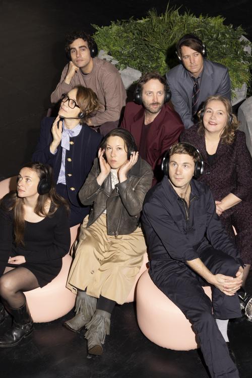 Das Ensemble von „Die Möwe“ sitzt verstreut auf beigen Sitzkissen. Sie tragen alle Overear-Kopfhörer und blicken in verschiedene Richtungen. Dabei haben sie unterschiedliche Gesichtsausdrücke: Manche lächeln, andere schauen gelangweilt und manche nachdenklich. Im Hintergrund steht ein großer Blumentopf mit gemischten Grünpflanzen.