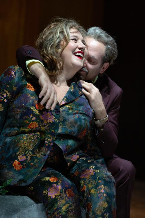 Bühnensituation. Bettina Stucky und Samuel Weiss sitzen auf einem grünen Sofa. Bettina trägt lockiges, schulterlanges Haar, roten Lippenstift und lacht und schaut an der Kamera vorbei. Sie trägt einen dunkelgrünen Samtanzug mit bunten Blumen-Applikationen drauf. Neben ihr sitzt Samuel, der einen Arm um ihre Schulter und die andere Hand auf ihre andere Schulter gelegt hat. Er dreht sein Gesicht leicht in ihre Haare, lächelt und hat die Augen geschlossen.