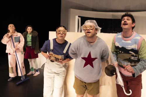 Das Ensemble steht vor großen weißen Bühnenbildteilen. Christine Ochsenhofer und Victoria Kraft stehen im Hintergrund links im Bild. Anastasia Lara Heller, Parsa Yaghoubi Pour und Silvio Kretschmer stehen vorne. Sie schauen erstaunt in die Ferne, Richtung Publikum. Parsa und Anastasia tragen beide eine große Brille aus Pappe. Anastasia hält eine Kamera in der Hand, sie ist ebenfalls aus Pappe. Silvio hält einen durchsichtigen Regenschirm und ein kleines Megafon in der Hand. 