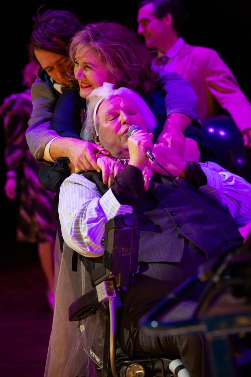 Bühnensituation. Josef Ostendorf sitzt im Vordergrund in einem Rollstuhl. Er trägt eine dunkle Anzugweste, ein helles Hemd, eine dunkle Hose und einen Brautschleier. In der rechten Hand hält er ein Mikro. Von hinten beugt sich Bettina Stucky über ihn und legt beide Arme um seinen Hals. Sie guckt nach rechts und lacht. Hinter Bettina steht Daniel Hoevels, der wiederum mit seinen Armen von hinten Bettinas Arme greift und sie umarmt. Ihre Gesichter sind in pinkes Licht getaucht. Im Hintergrund ist Pascal Houdu
