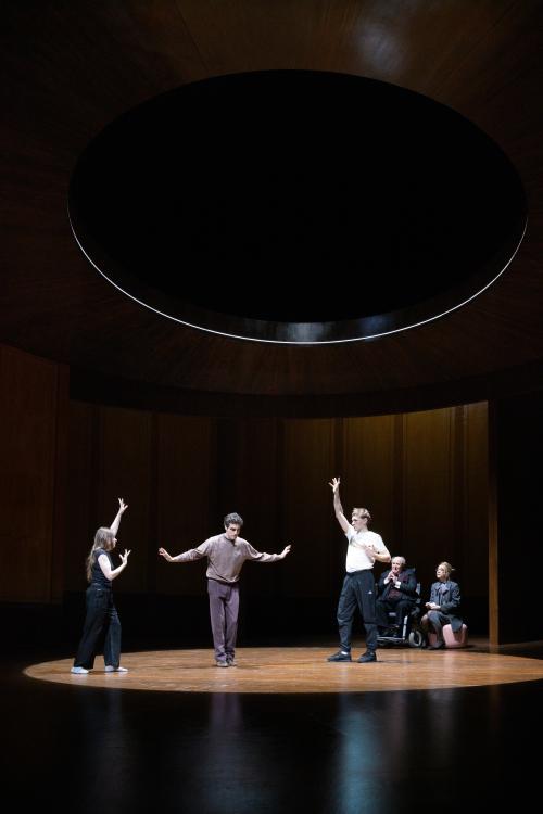 Bühnensituation. Henni Jörissen, Pascal Houdus und Paul Behren stehen in einem holzgetäfelten Raum. Sie haben die Arme erhoben und tanzen. Ein runder Kegel aus Licht ist nur auf sie gerichtet. Am Rand des Lichtkegels sitzen Angelika Richter auf einem rosafarbenen Pouf und Josef Ostendorf in einem Rollstuhl. Beide betrachten die Tanzenden.