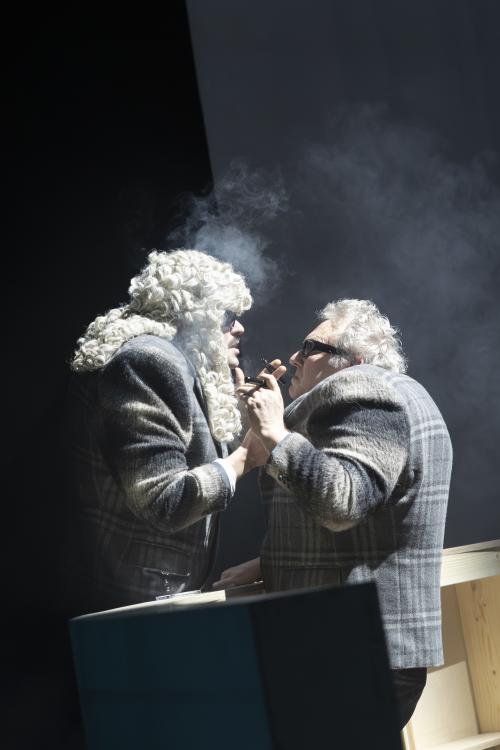 Parsa Yaghoubi Pour und Hermann Book stehen sich gegenüber und schauen sich ins Gesicht. Zwischen ihnen ein Bühnenbildteil, dass ihnen ungefähr bis zur Hüfte reicht. Die Umgebung ist dunkel, nur die beiden Schauspieler stehen im Licht. Parsa trägt eine große, graue Perücke mit Locken. Beide tragen dunkle Sonnenbrillen und den gleichen Anzug, der grau-kariert ist. In ihren Händen halten sie Zigarren-artige Gegenstände, die qualmen. 