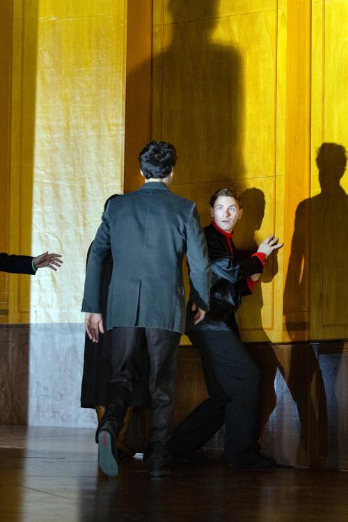 Bühnensituation. Paul Behren steht in einem großen, holzvertäfelten Raum und trägt einen schwarzen Anzug mit roten Akzenten. Sein Körper ist zur Wand gewannt, doch er schaut ins Publikum. Pascal Houdus geht auf ihn zu, mit dem Rücken zum Publikum. Er trägt einen schwarzen Anzug.