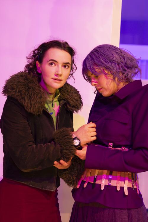 Victoria Kraft und Anastasia Lara Heller stehen sich gegenüber. Während Victoria mit aufgerissenen Augen zur Seite starrt, blickt Anastasia nach unten auf ihre Hände, die an Victorias Armbanduhr fassen. Victoria trägt eine dunkelbraune Felljacke und Anastasia eine lilafarbene Perücke und über eine lilafarbene Anzugjacke einen Gürtel bestehend aus Armbanduhren. 