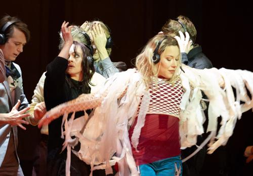 Josefine Israel trägt ein rotes Oberteil und darüber ein weißes Netzoberteil, das an den Armen mit langen und großen Fransen versehen ist. Sie bewegt sich mit ausgestreckten Armen tänzerisch. Im Hintergrund ist das Ensemble zu sehen, sie alle tragen schwarze Kopfhörer auf dem Kopf und bewegen ihre Hände und Arme. 