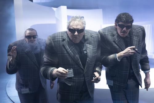 Christine Ochsenhofer, Hermann Book und Silvio Kretschmer stehen nebeneinander in einer Reihe. Unter ihren karierten Anzügen tragen sie sehr breite Schulterpolster. Alle drei haben Sonnenbrillen auf und Zigarren in den Händen. Hermann in der Mitte hält einen Ausweis in der Hand.