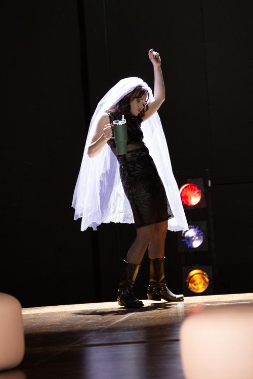 Henni Jörissen bewegt sich mit geschlossenen Augen. Sie trägt ein schwarzes Kleid, schwarze Biker-Stiefel und einen langen Brautschleier auf dem Kopf. Sie hält einen Trinkbecher in der einen Hand, die andere streckt sie nach oben.