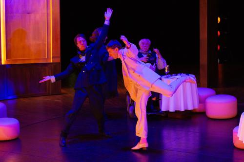 Paul Behren trägt einen weißen und Daniel Hoevels einen schwarzen Anzug. Sie bewegen sich in einem holzgetäfelten Raum mit erhobenen Armen und tanzen. Das Licht wirft bunte Farben auf die Bühne. Josef Ostendorf und Bettina Stucky sind in Hintergrund zu sehen. Josef sitzt in einem Rollstuhl und raucht. Neben ihm steht ein gedeckter Tisch mit weißer Tischdecke.