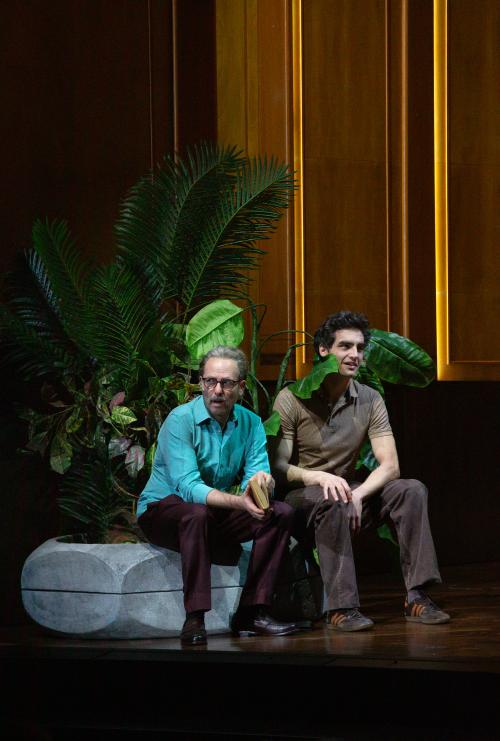 Samuel Weiss und Pascal Houdus sitzen in einem großen, holzvertäfelten Raum auf dem Rand eines großen Blumentopfes, aus dem palmenartigen Blätter herausragen. Eins der Blätter berührt Pascal Houdus Schulter. Samuel trägt ein türkises Polohemd und eine braune Hose und Pascal ein braunes Polohemd und eine braune Hose. Samuel hält ein Buch in den Händen und blickt zur Seite.