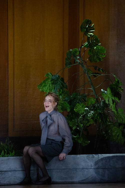 Angelika Richter sitzt in einem großen, holzvertäfelten Raum auf dem Rand eines großen Blumentopfes, aus dem palmenartigen Blätter herausragen. Sie trägt eine Brille, eine graue Bluse mit einer Schleife vorne dran und einen schwarzen Rock und Strumpfhose. Sie lacht mit offenem Mund.