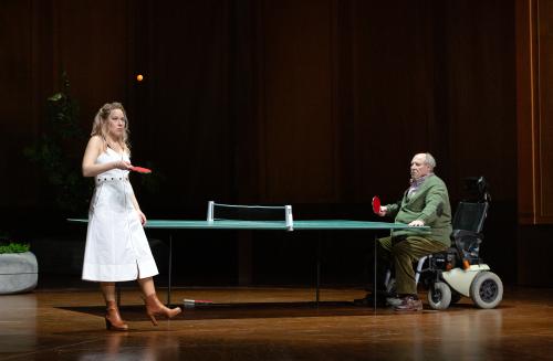 Bühnensituation. Josefine Israel steht neben einer grünen Tischtennisplatte. Sie trägt braune Lederstiefel mit Absätzen und ein weißes schulterfreies Kleid. Sie hält einen Tischtennisschläger in der rechten Hand und lässt einen Ball darauf springen. Ihr Blick ist auf den Ball in der Luft gerichtet. Am anderen Ende der Tischtennisplatte sitzt Josef Ostendorf in einem Rollstuhl. Er trägt eine grüne Hose, braune Halbschuhe und eine grüne Strickjacke über einem Hemd. In der rechten Hand hält er einen Tischtenni