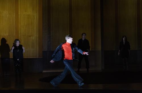 Bühnensituation. Paul Behren ist im Vordergrund, er geht mit großen Schritten über die Bühne.  Er trägt einen schwarzen Anzug mit einem roten Oberteil darunter. Im Hintergrund stehen Bettina Stucky, Daniel Hoevels und Henni Jörissen, die in schwarz gekleidet sind. 