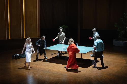 Bühnensituation. Josefine Israel, Henni Jörissen, Pascal Houdus, Daniel Hoevels, Samuel Weiss und Bettina Stucky haben alle Tischtennisschläger in den Händen und laufen um eine Tischtennisplatte herum. Im Hintergrund sind holzvertäfelte hohe Wände zu sehen. Auch der Boden ist als Holz. Etwas abseits liegt Paul Behren auf dem Boden.