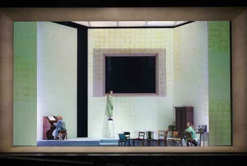 Bühnensituation: Das Bühnenbild besteht aus hohen weißen Wänden auf denen grüne überlappende Kreise aufgemalt sind. Die mittlere Wand hat eine große rechteckige Öffnung, dahinter ist es schwarz. Ilse Ritter sitzt mit dem Rücken zum Publikum links an einem Klavier. Mittig steht Alberta von Polnitz auf einer Leiter. Rechts sitzt Julia Wieninger auf einem Stuhl. Alle drei tragen grün-blaue Bademäntel. Mehrere Stühle sind auf der Bühne verteilt. Rechts ein Holzschrank sowie ein Tonbandgerät auf einem Tisch.