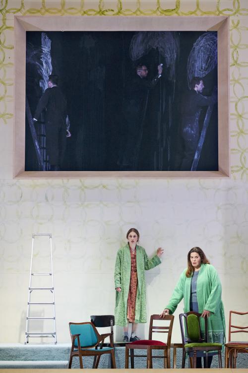 Bühnensituation: Alberta von Polnitz und Julia Wieninger schauen geradeaus. Sie tragen beide einen grünen Bademantel. Vor ihnen sind mehrere Stühle verteilt, links steht eine Leiter. Im Hintergrund eine weiße Wand mit aufgemalten grünen Kreisen. Die Wand hat eine große rechteckige Öffnung. In der Öffnung ist es dunkel, es sind drei Männer in schwarzen Anzügen auf Leitern zu sehen, die mit weißer Kreide die schwarze Wand bemalen. 