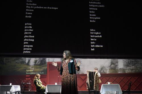 Bühnensituation: Julia Wieninger steht im Vordergrund und schaut zur Seite, die Hände erhoben. Hinter ihr drei Tische mit weißen Tischdecken und weißen Bechern darauf. Alberta von Polnitz sitzt an dem mittleren. Über ihnen eine schwarze Leinwand, auf die weißer Worte untereinander projiziert werden: fische, fischreiher, flug der gedanken aus dem körper, flüstern, frauen, friede, gehirn, geiseln, gewehre …