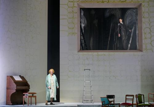 Bühnensituation: Ilse Ritter steht in einem grün-blauen Bademantel auf der Bühne und lächelt. Links von ihr ein Klavier aus Holz. Rechts von ihr eine Leiter und mehrere Stühle. Im Hintergrund eine hohe weiße Wand mit aufgemalten grünen Kreisen. Die Wand hat eine große rechteckige Öffnung. In der Öffnung ist es dunkel, ein Mann in einem schwarzen Anzug auf einer Leiter ist zu sehen, der zu ihr Ilse Ritter schaut. Die schwarze Wand hinter ihm ist mit verwischter Kreide bemalt.