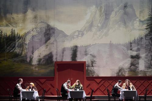 Bühnensituation: Ilse Ritter, Alberta von Polnitz und Julia Wieninger sitzen an drei kleinen Tischen mit weißen Tischdecken und weißen Bechern darauf. Mit ihnen am Tisch jeweils ein Mann in weißem Hemd, schwarzer Fliege und schwarzer Weste. Im Hintergrund ist eine große Wandmalerei von einer Berglandschaft mit Tannen zu sehen. 