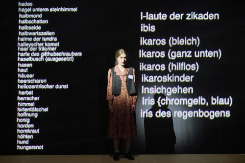 Bühnensituation: Alberta von Polnitz steht in einem gemusterten orangefarbenen Kleid mit schwarzer Weste auf der Bühne und schaut geradeaus. Links und rechts von ihr werden auf eine schwarze Wand untereinander weiße Worte projiziert: halbmond, halbschatten, halbseide, halbwertszeiten ….