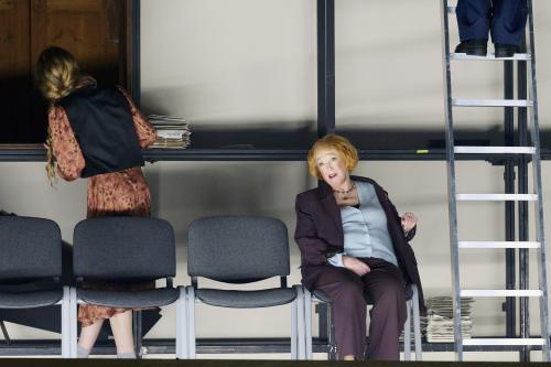 Bühnensituation: Ilse Ritter sitzt auf einem Stuhl. Neben ihr in einer Reihe weitere Stühle. Im Hintergrund steht ein großes Metallgestell, an dem Alberta von Polnitz mit dem Rücken zu ihr steht. Rechts steht eine Leiter, es sind zwei Schuhe und blaue Hosenbeine zu sehen.