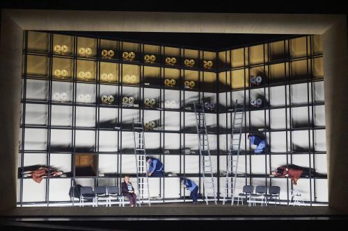 Bühnensituation: Über das Bühnenbild erstreckt sich ein Metallgestell, welches so angeordnet ist, dass sich regalartige Fächer bilden. In den oberen Fächern sind mehrere Tonbandgeräte zu sehen. Etwas weiter unten knien verteilt drei Männer in einzelnen Regalfächern.  Alberta von Polnitz und Julia Wieninger liegen jeweils links und rechts in Fächern, Ilse Ritter sitzt auf einem Stuhl in einer Stuhlreihe.