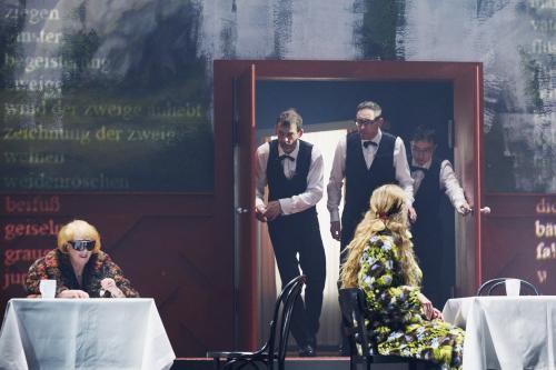 Bühnensituation: Ilse Ritter sitzt an einem Tisch mit einer weißen Tischdecke und einem weißen Becher darauf. Sie trägt eine große Sonnenbrille. Neben ihr Alberta von Polnitz an einem weiteren, identischen Tisch. Im Hintergrund ist eine große Wandmalerei von einer Berglandschaft mit Tannen zu sehen. Inmitten befindet sich eine rote Tür, aus der drei Männer mit schwarzen Hosen, weißen Hemden, schwarzer Weste und Fliege herauskommen, sie schauen zu Ilse Ritter. 