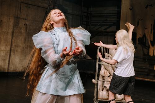 Im Vordergrund steht Christiane von Poelnitz, die Hände geöffnet nach vorn ausgestreckt. Ihre Augen sind geschlossen, ihr Mund ist offen und zu einem Lächeln gezogen, sie streckt das Gesicht nach oben. Im Hintergrund steht Henni Jörissen abgewendet. Sie streckt ihre beiden Armen nach vorn. Hinter ihr steht ein Rollwagen mit auseinander gebauten Schaufensterpuppen.