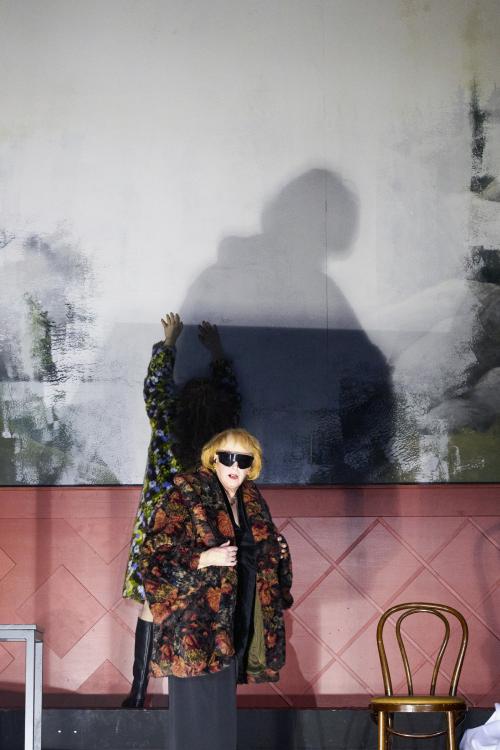 Bühnensituation: Ilse Ritter steht im Vordergrund. Sie trägt eine geblümte Jacke, eine große Sonnenbrille und schaut ins Publikum. Neben ihr ein leerer Stuhl.