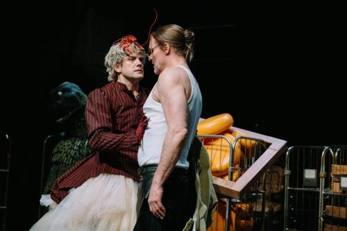 Christoph Jöde und Yorck Dippe stehen sich gegenüber. Yorck Dippe trägt ein weißes Muskelshirt und eine schwarze Hose, seine blonden Haare sind zu einem Dutt nach hinten gesteckt. Christoph Jöde steht vor ihm und blickt ihn an. Auf seinen blonden Locken sitzt ein Plastikhummer. Seine Hand im roten Handschuh liegt auf Yorck Dippes Oberkörper. Er trägt ein schwarz-rot gestreiftes Jackett mit Puffärmeln sowie einen Rock aus Tüll. Im Hintergrund sieht man Rollwägen, in einem sind Käselaibe aufgetürmt.