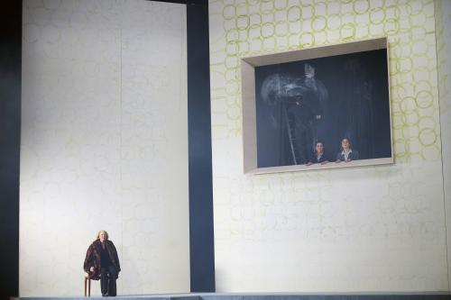 Bühnensituation: Ilse Ritter sitzt auf einem Stuhl. Im Hintergrund zwei hohe weiße Wände mit aufgemalten grünen Kreisen. Eine Wand hat eine große rechteckige Öffnung. Aus der Öffnung gucken Alberta von Polnitz und Julia Wieninger heraus. Hinter ihnen ein Mann im schwarzen Anzug auf einer Leiter, dahinter verwischte Kreidespuren an der Wand.