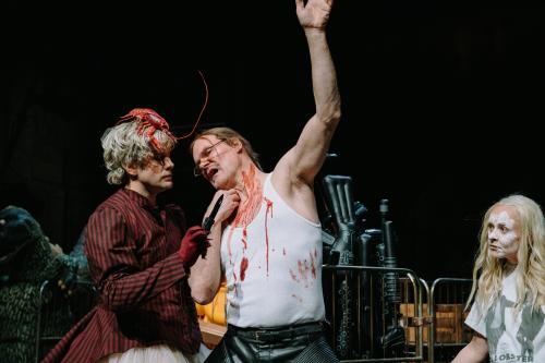 Yorck Dippe trägt ein weißes Muskelshirt und eine schwarze Lederhose. Rote Farbe läuft seinen Hals herunter auf sein weißes Oberteil. Christoph Jöde steht vor ihm, hält ein schwarzes Messer in der Hand, das er anschaut. Auf seinen blonden Locken sitzt ein Plastikhummer. Seine Hand steckt in einem roten Handschuh. Er trägt ein schwarz-rot gestreiftes, zugeknöpftes Jackett mit Puffärmeln. Rechts im Bild steht Henni Jörissen mit langen Haaren und weißer Farbe im Gesicht. Sie schaut zu den beiden Männern. 