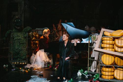 Christoph Jöde sitzt im Tüllrock und schwarz-rot gestreiften Jackett mit Puffärmeln am schwarzen Flügel. Christiane von Poelnitz sitzt davor und hat die Augen geschlossen, sie ist ganz in schwarz gekleidet. Über die Bühne sind diverse vergitterte Rollwägen verteilt mit verschiedener Beladung: Ein grünes, Dinosaurier-ähnliches Monster, goldene Schaufensterpuppenköpfe, Schweinehälften und Käselaibe. Auf dem Boden liegen verstreut Blumen.