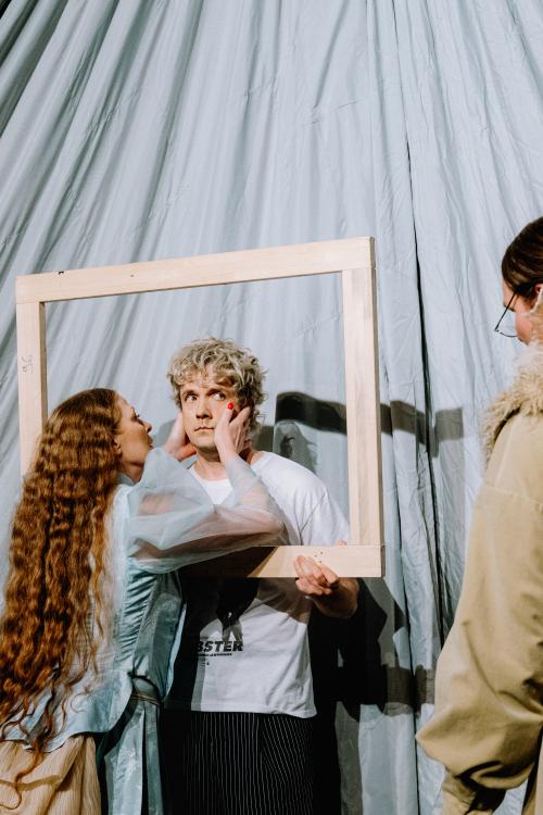 Christiane von Poelnitz, Christoph Jöde und Yorck Dippe stehen vor einem hellen Hintergrund aus Stoff. Christiane von Poelnitz trägt ein blaues Rüschenoberteil mit Puffärmel aus Chiffon. Sie hat die Hände an Christoph Jödes Gesicht gelegt und schaut ihn an. Christoph Jöde hat blonde Locken, trägt ein weißes T-Shirt mit schwarzem Print und eine schwarze Hose mit weißen Streifen. Er hält einen Holzrahmen in den Händen, durch den er schaut. Sein Blick geht zu Yorck Dippe, rechts im Bild abgeschnitten ist.