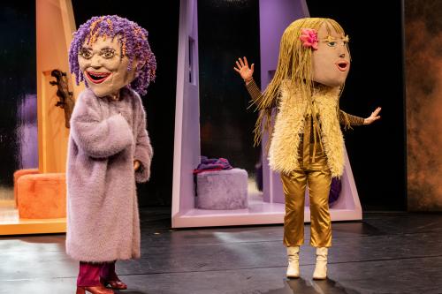 Bühnensituation: Zwei Personen mit aufgesetzten großen Kopfmasken nebeneinander. Die linke Person trägt rote Stiefel, eine rote Hose, einen lilafarbenen flauschigen Mantel, ihre Kopfmaske hat lilafarbene Locken mit orangenen Strähnen und einen geöffneten, lächelnden Mund. Die rechte Person trägt goldene Kleidung zu weißen Stiefeln und eine helle Fellweste, ihre Kopfmaske hat rückenlange blonde Haare mit einer rosa Schleife. Der Mund ist geöffnet. Im Hintergrund angedeutete orange- und lilafarbene Räume.
