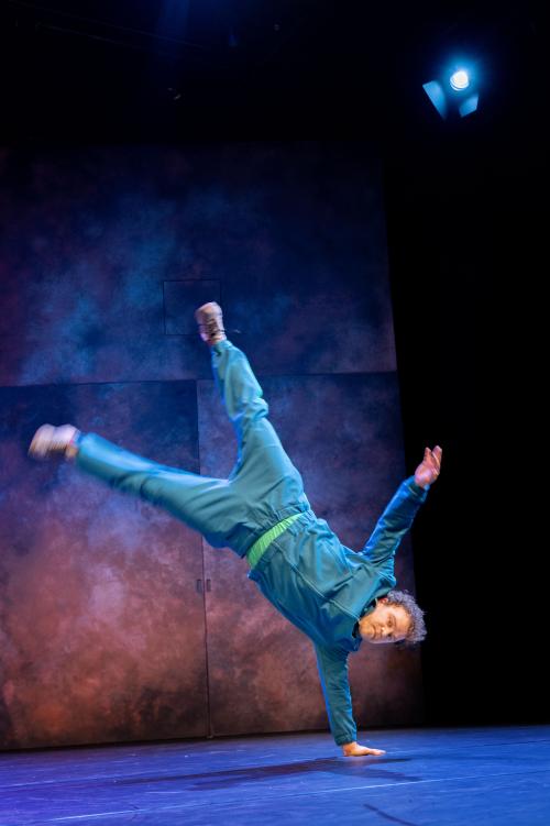 Bühnensituation: Parsa Yaghoubi Pour in einem blauen Trainingsanzug stützt sich mit einer Hand am Boden ab, der restliche Körper befindet sich in der Luft, seine Beine sind ausgestreckt.