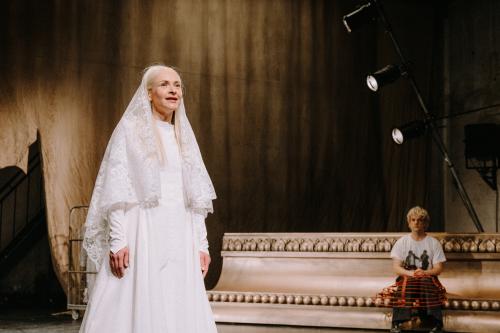 Links im Vordergrund steht Henni Jörissen in einem langen, weißen Kleid mit spitzzulaufenden Ärmeln, sie trägt einen Schleier aus Spitze auf dem Kopf und hat den Mund geöffnet. Der Hintergrund ist braun und beige. Im Hintergrund liegt ein überdimensional großer, unterer Teil eines Bilderrahmens im Barockstil in goldenem Farbton. Auf ihm sitzt Christoph Jöde, seine Hände auf seinem Schoß. Er trägt ein weißes T-Shirt. Über seiner schwarzen Hose trägt er einen roten Reifenrock. Er blickt zu Henni Jörissen. 