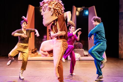 Bühnensituation: Das Ensemble trägt bunte, einfarbige Kleidung in lila, gold, blau und lila. Alle tanzen. Eine Person trägt eine übergroße Kopfmaske mit Gesicht und Haaren. Im Hintergrund kleine, einfarbige hohe Räume in orange und lila.