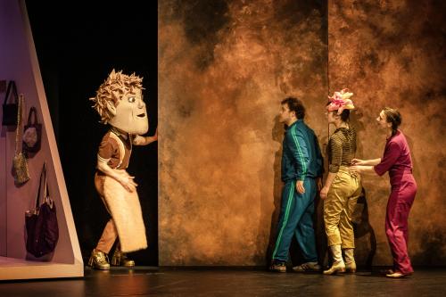 Bühnensituation: Eine Person mit braun-rosa Kleidung und einer großen Kopfmaske mit Gesicht hält sich an einer goldenen Wand fest. Sie schaut in Richtung von Parsa Yaghoubi Pour, Anastasia Lara Heller und Viktoria Kraft, die hintereinander an der gleichen Wand kurz vor der Person mit Maske stehen. 