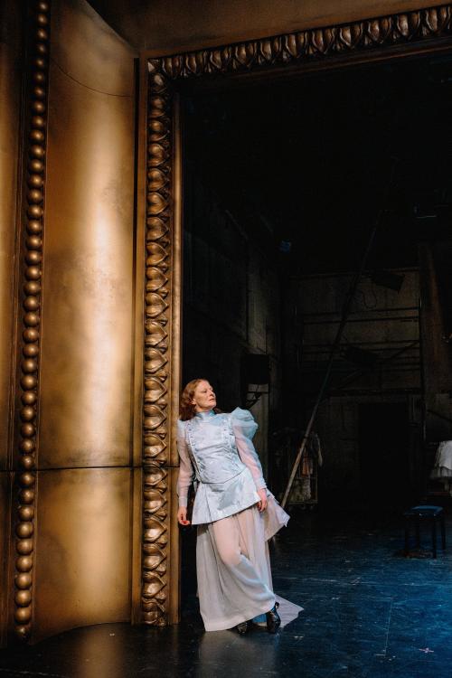 Christiane von Poelnitz in blauem Rüschenoberteil mit Puffärmel aus Chiffon und einen beigen Rüschenrock lehnt seitlich an einem überdimensional großen, goldenen, verzierten Barockbilderrahmen. Sie steht auf ihrem linken Bein, das rechte Bein hat sie über das linke geschlagen. 