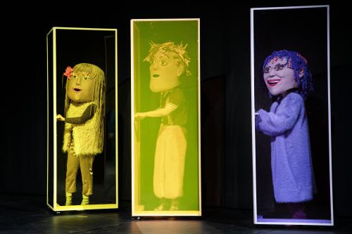 Bühnensituation: Drei Personen mit großen Kopfmasken stehen einzeln in drei hohen, eckigen Kästen. Alle haben die Hand auf einer Türklinke liegen.