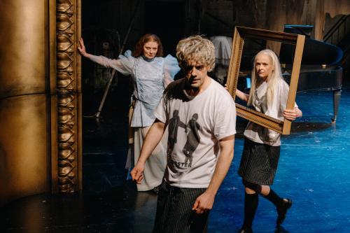 Im Vordergrund steht Christoph Jöde, sein Mund ist leicht geöffnet, seine Augenbrauen zusammengezogen. Christiane von Poelnitz steht links dahinter, stützt sich an den großen Bilderrahmen im Barockstil. Sie schaut Christoph Jöde an. Henni Jörissen steht sich rechts hinter Christoph Jöde, trägt einen Bilderrahmen vor sich und wie Christoph Jöde ein weißes Oberteil. Dazu trägt sie eine schwarz-weiße Shorts, schwarze Kniestrümpfe und Schuhe. Sie sieht Christoph Jöde an, ihre Stirn ist in Falten gelegt. 