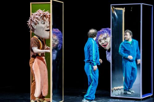 Bühnensituation: Links eine Person in braun-rosafarbener Kleidung mit einer großen Kopfmaske mit braun-rosa Haaren. Sie steht im Eingang eines hohen schmalen Kastens mit verspiegelten Wänden. Mittig steht Parsa Yaghoubi Pour in einem blauen Trainingsanzug, der sich zu einer Person mit einer weiteren Kopfmaske dreht, die lilafarbene Locken mit orangenen Strähnen und einen geöffneten, lächelnden Mund hat. Sie lehnt sich hinter einem verspiegelten hohen Kasten hervor. In diesem spiegelt sich Parsa.