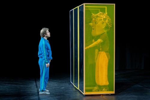 Bühnensituation: Parsa Yaghoubi Pour in einem blauen Trainingsanzug, steht vor drei nebeneinanderstehenden hohen eckigen Kästen. Die Seitenwand ist grünlich transparent. In einem Kasten steht eine Person in braun-rosafarbener Kleidung mit einer großen Kopfmaske mit braun-rosa Haaren mit der Hand an einer Türklinke. Parsa schaut sie an. 