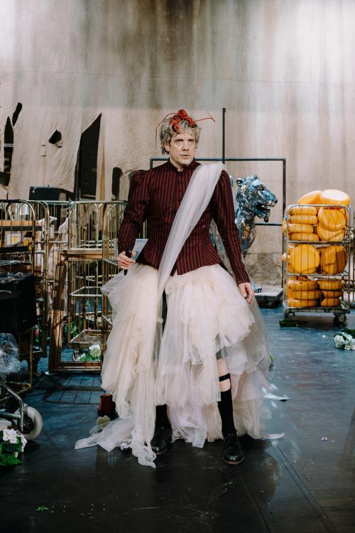 Christoph Jöde steht der Kamera zugewandt aufrecht da. Er trägt einen Rock und eine Schärpe aus Tüll, schwarze Kniestrümpfe und schwarze Schuhe. Sein Oberteil ist ein schwarz-rot gestreiftes, zugeknöpftes Jackett mit Puffärmeln. Auf seinen blonden Locken trägt er einen Plastikhummer. In seiner linken Hand hält er ein schwarzes Messer. Im Hintergrund sind diverse vergitterte Rollwägen, in einem sind unzählige Käselaibe. Die hintere Stoffwand hat Risse.